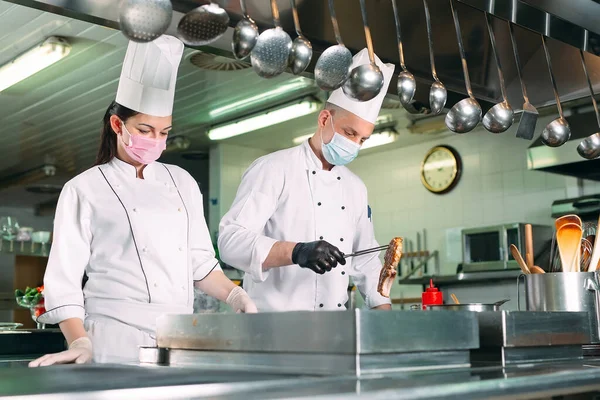 Chefs con máscaras protectoras y guantes preparan comida en la cocina de un restaurante u hotel. —  Fotos de Stock