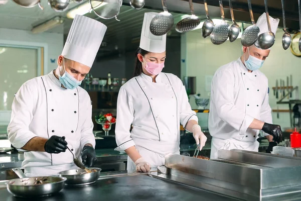 Chefs em máscaras e luvas de proteção preparar alimentos na cozinha de um restaurante ou hotel. — Fotografia de Stock