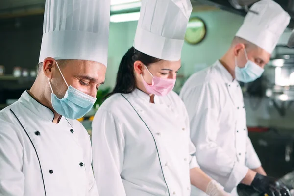 Chefs con máscaras protectoras y guantes preparan comida en la cocina de un restaurante u hotel. —  Fotos de Stock