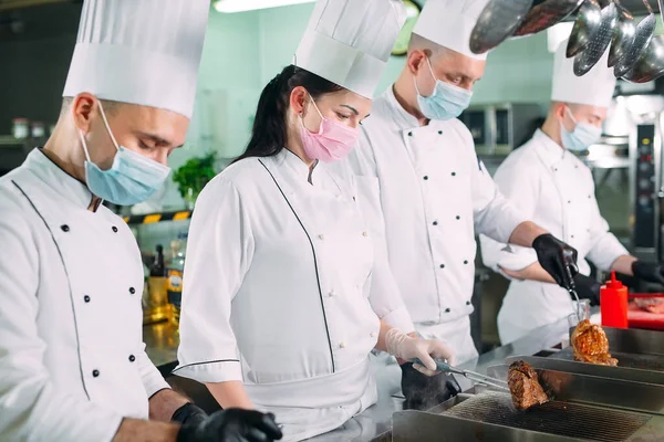 Chefs con máscaras protectoras y guantes preparan comida en la cocina de un restaurante u hotel. — Foto de Stock