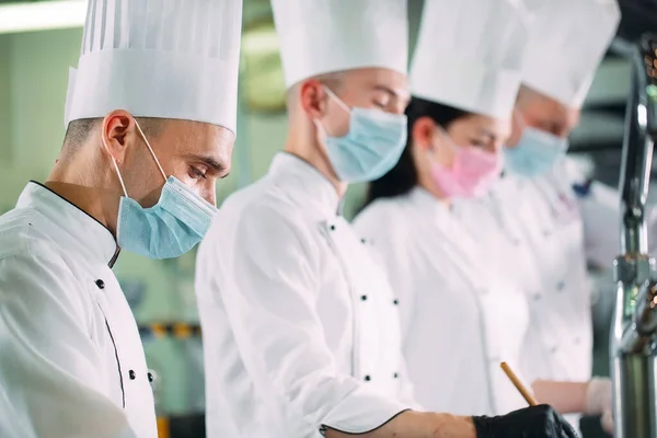 Кухарі в захисних масках і рукавичках готують їжу на кухні ресторану або готелю . — стокове фото