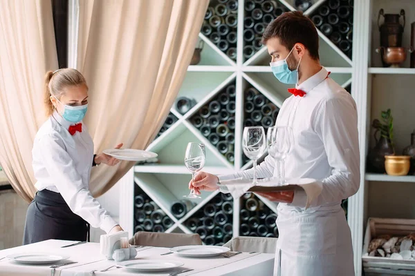 Ένας σερβιτόρος με ιατρική προστατευτική μάσκα σερβίρει ένα τραπέζι στο εστιατόριο. Υπάλληλοι εστιατορίου ή ξενοδοχείου με προστατευτικές μάσκες. Το τέλος της καραντίνας. — Φωτογραφία Αρχείου