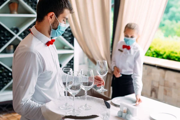 Un cameriere con una maschera protettiva medica serve un tavolo nel ristorante. Dipendenti di un ristorante o di un hotel con maschere protettive. La fine della quarantena. — Foto Stock