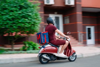 Scooter 'ın üzerinde kırmızı üniformalı teslimatçı çocuk ve hızlı giden izotermal yiyecek kutusu. Kafelerden ve restoranlardan ekspres yemek servisi. Moto scooter 'daki kurye yiyecek getiriyor..