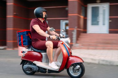 Scooter 'ın üzerinde kırmızı üniformalı teslimatçı çocuk ve hızlı giden izotermal yiyecek kutusu. Kafelerden ve restoranlardan ekspres yemek servisi. Moto scooter 'daki kurye yiyecek getiriyor..