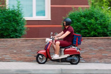 Scooter 'ın üzerinde kırmızı üniformalı teslimatçı çocuk ve hızlı giden izotermal yiyecek kutusu. Kafelerden ve restoranlardan ekspres yemek servisi. Moto scooter 'daki kurye yiyecek getiriyor..