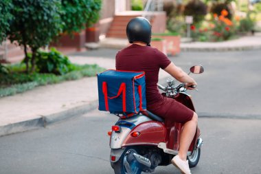 Scooter 'ın üzerinde kırmızı üniformalı teslimatçı çocuk ve hızlı giden izotermal yiyecek kutusu. Kafelerden ve restoranlardan ekspres yemek servisi. Moto scooter 'daki kurye yiyecek getiriyor..