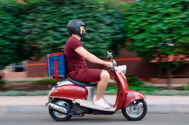 Scooter 'ın üzerinde kırmızı üniformalı teslimatçı çocuk ve hızlı giden izotermal yiyecek kutusu. Kafelerden ve restoranlardan ekspres yemek servisi. Moto scooter 'daki kurye yiyecek getiriyor..