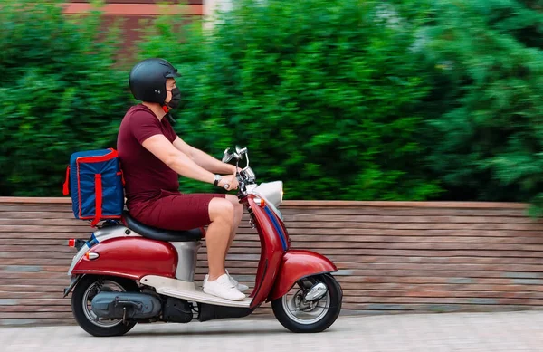 Scooter 'ın üzerinde kırmızı üniformalı teslimatçı çocuk ve hızlı giden izotermal yiyecek kutusu. Kafelerden ve restoranlardan ekspres yemek servisi. Moto scooter 'daki kurye yiyecek getiriyor..