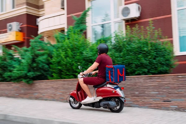 Entrega niño vistiendo uniforme rojo en scooter con caja de comida isotérmica conduciendo rápido. Servicio de entrega urgente de comida desde cafeterías y restaurantes. Mensajero en la moto scooter entrega de alimentos. — Foto de Stock