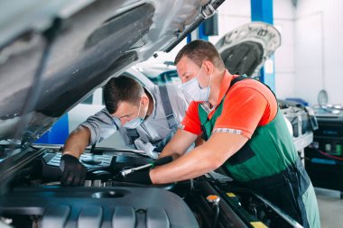 Araba atölyesinde üniformalı ve koruyucu maskeli tamirciler çalışıyor..