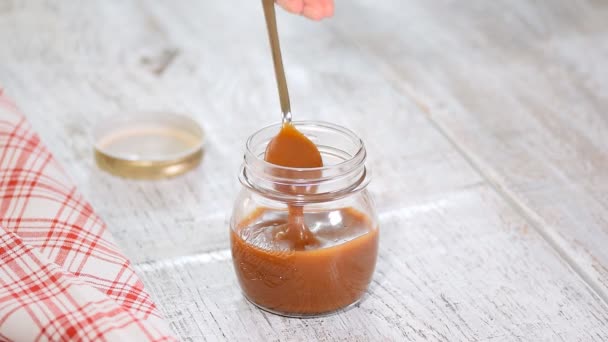 Cucchiaio con gustosa salsa al caramello sopra il barattolo sul tavolo . — Video Stock