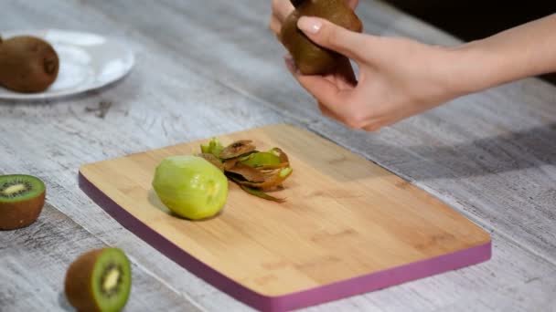 Een vrouw reinigt de schil van de kiwi met een mes op een houten achtergrond. — Stockvideo