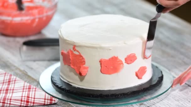 Mão de confeiteiro esfrega creme rosa no bolo de creme de esponja de gelo branco na plataforma giratória de madeira na cozinha da padaria, de perto . — Vídeo de Stock