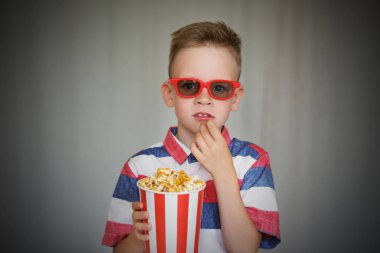 Genç çocuk içinde 3d gözlük sinemada ya da evde bir film izlemek. Küçük çocuk gri arka plan üzerinde patlamış mısır yemek. Ev sineması. Vintage sinema gözlük şirin çocuk. Eğlence kavramının. 
