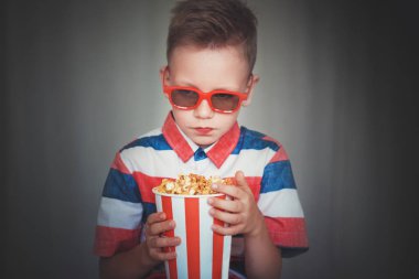 Genç çocuk içinde 3d gözlük sinemada ya da evde bir film izlemek. Küçük çocuk gri arka plan üzerinde patlamış mısır yemek. Ev sineması. Vintage sinema gözlük şirin çocuk. Eğlence kavramının. 
