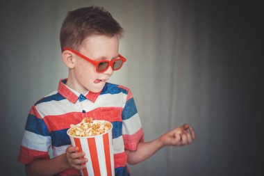 Genç çocuk içinde 3d gözlük sinemada ya da evde bir film izlemek. Küçük çocuk gri arka plan üzerinde patlamış mısır yemek. Ev sineması. Vintage sinema gözlük şirin çocuk. Eğlence kavramının. 