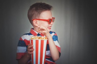 Genç çocuk içinde 3d gözlük sinemada ya da evde bir film izlemek. Küçük çocuk gri arka plan üzerinde patlamış mısır yemek. Ev sineması. Vintage sinema gözlük şirin çocuk. Eğlence kavramının. 