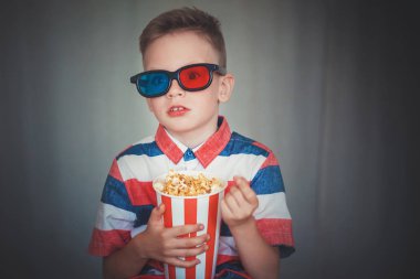 Genç çocuk içinde 3d gözlük sinemada ya da evde bir film izlemek. Küçük çocuk gri arka plan üzerinde patlamış mısır yemek. Ev sineması. Vintage sinema gözlük şirin çocuk. Eğlence kavramının. 