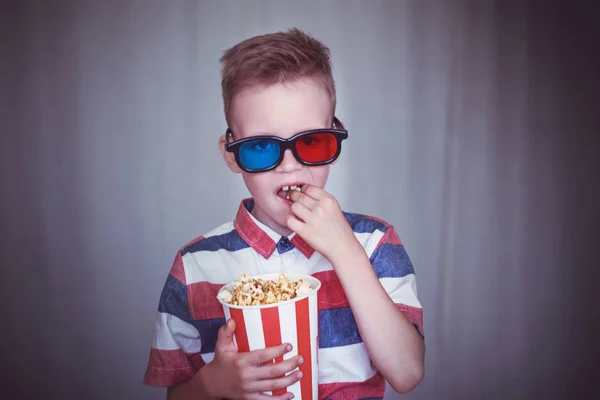 Young boy watch a movie in 3D glasses at the cinema or at home. Little kid eat popcorn over gray background. Home theater. Cute Child in vintage cinema eyeglasses. Entertainment concept.