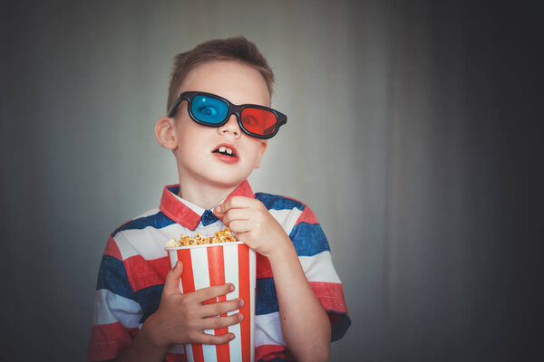 Young boy watch a movie in 3D glasses at the cinema or at home. Little kid eat popcorn over gray background. Home theater. Cute Child in vintage cinema eyeglasses. Entertainment concept. 