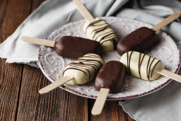 Milk chocolate popsicles on a stick. Ice cream popsicles covered with white and dark chocolate on the plate on wooden background. Space for text. Chocolate ice cream bars, nuts. Ice cream dessert