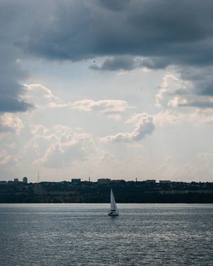Dramatik ve bulutlu bir gökyüzünün altında sakince yüzen bir yelkenli manzarası. Şehrin ufuk çizgisi arka planda görünüyor, doğa ve medeniyet arasında bir tezat oluşturuyor..