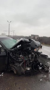 Islak bir yolda kaza yaptıktan sonra kaza yapmış siyah bir araba. Ön tarafı parçalanmış ve kaputu açık bir araba şehir caddesinde. Yağmurlu hava.