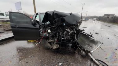 Islak bir yolda kaza yaptıktan sonra kaza yapmış siyah bir araba. Ön tarafı parçalanmış ve kaputu açık bir araba şehir caddesinde. Yağmurlu hava