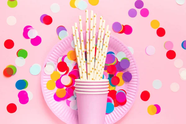 Birthday or party paper glasses with straws, dish and colorful confetti on pink background. Festive concept. Flat lay style. Top view.