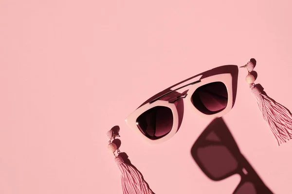 Composition made with woman accessories. Sunglasses and earrings on pink background. Creative monochrome layout. Fashion and summer concept.