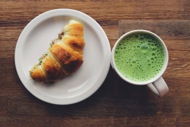 Kafedeki ahşap bir masada beyaz bir tabakta bir fincan kibritli latte ve kruvasan. Üst görünüm.