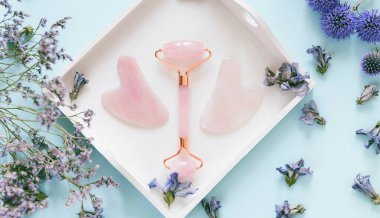 Ev spası için yüz masajı seti. Gül kuvarstan yapılmış yüz silindiri ve gua sha masaj aletleri pastel mavi arka planda beyaz ahşap tepside çiçekli. Doğal tedavi kavramı.