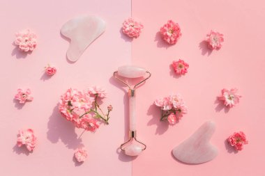Ev spası için yüz masajı seti. Gül tomurcuklarıyla pembe pastel arka planda gül kuvarsından yapılmış yüz silindiri ve gua sha masaj aleti. Doğal tedavi kavramı.