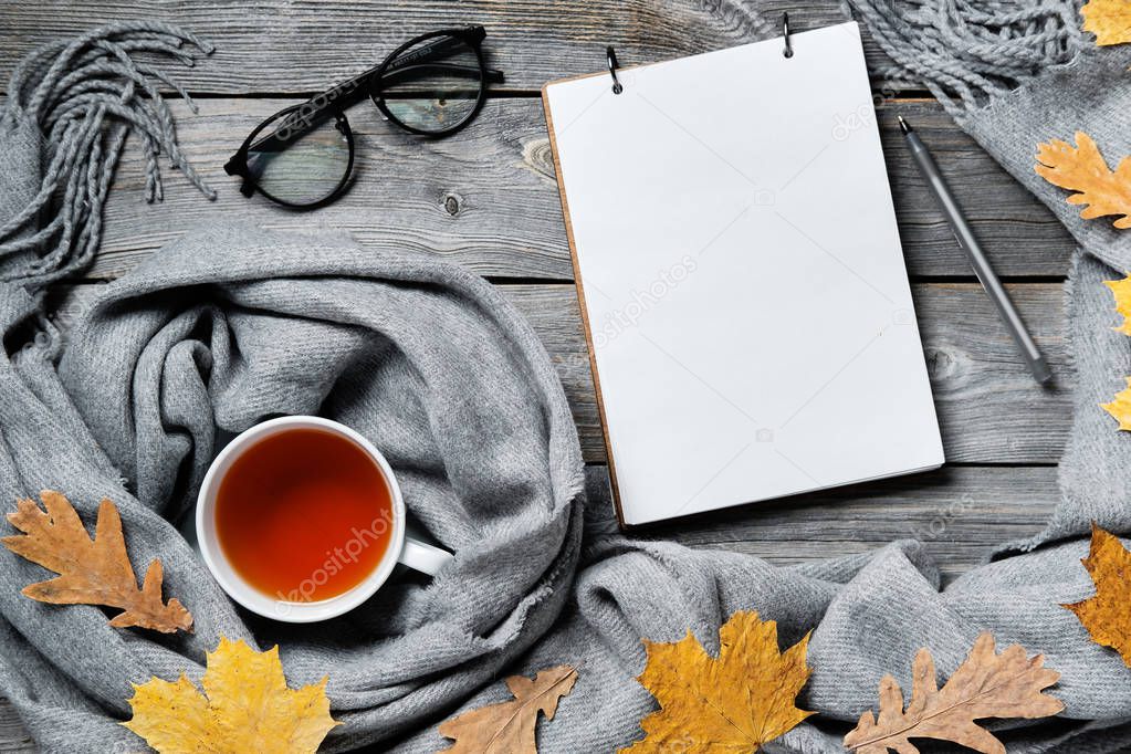 Autumn composition, fall leaves, hot steaming cup of tea and a warm scarf on wooden table background. Notepad with pen