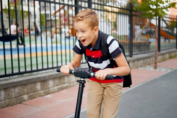 De jongen van de school in rijden met zijn elektrische scooter in de stad met rugzak op zonnige dag. Kind in kleurrijke kleding fietsen op weg naar school — Stockfoto