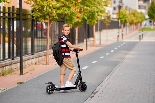 De jongen van de school in rijden met zijn elektrische scooter in de stad met rugzak op zonnige dag. Kind in kleurrijke kleding fietsen op weg naar school — Stockfoto