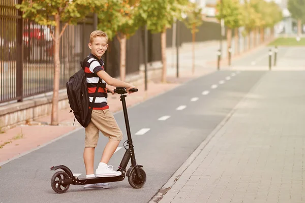 Menino da escola em andar com sua scooter elétrica na cidade com mochila no dia ensolarado. Criança em roupas coloridas de bicicleta a caminho da escola — Fotografia de Stock