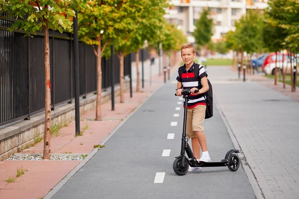 Menino da escola em andar com sua scooter elétrica na cidade com mochila no dia ensolarado. Criança em roupas coloridas de bicicleta a caminho da escola — Fotografia de Stock