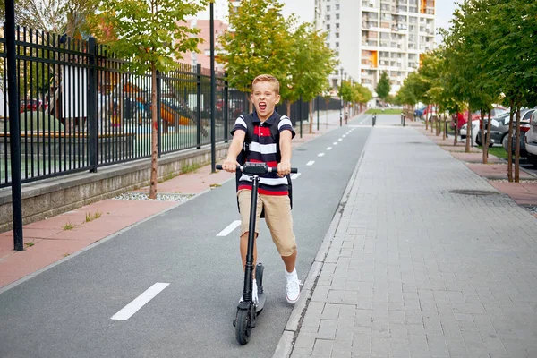 De jongen van de school in rijden met zijn elektrische scooter in de stad met rugzak op zonnige dag. Kind in kleurrijke kleding fietsen op weg naar school — Stockfoto