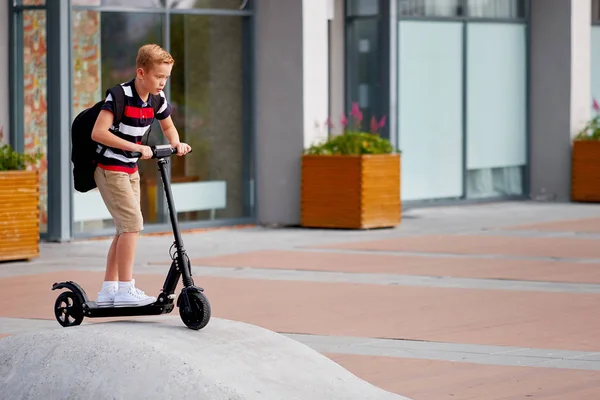 Menino da escola em andar com sua scooter elétrica na cidade com mochila no dia ensolarado. Criança em roupas coloridas de bicicleta a caminho da escola — Fotografia de Stock