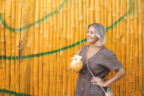 Young Woman on Vacation Drink Coconut Juice. Portrait of Happy Lady Hold Exotic Fresh Cocktail on Yellow Bamboo Stick Background. Female Tourist Smile Relax on Summer Holiday