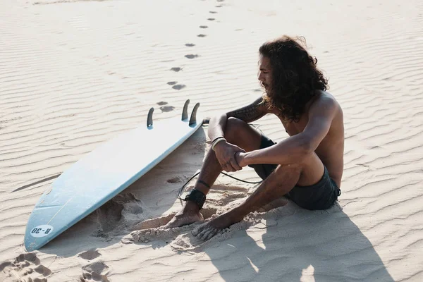 Hombre Surfista sentado en la tabla de surf en la playa de arena —  Fotos de Stock