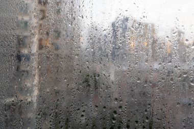 Yoğunlaşma ve yağmur damlalarıyla kaplı cam bir pencere. Yüksek binaların belirsiz çizgileri ve parlayan şehir ışıkları.