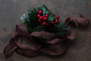 Parlak kırmızı böğürtlenlerle süslenmiş şenlikli çelenk, yapay çam filizleri sahte kar ve yeşil yapraklarla kaplanmış. Kırsal bir kahverengi kurdele düzenlemenin etrafına ustalıkla örtülmüş, karanlık bir metne karşı basit ama büyüleyici bir Noel süsü oluşturuyor.