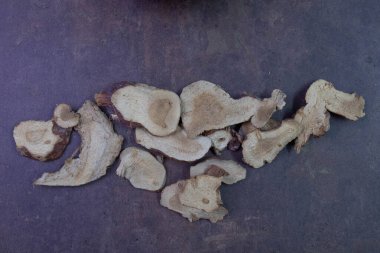 Galangal dilimleriyle dolu siyah bir kase. Ek parçalar koyu dokulu bir yüzeye saçılmış. Topraksı tonlar ve organik şekiller, geleneksel aşçılık ya da yemek pişirme gibi ilaçlarda sıklıkla kullanılan kurutulmuş otların doğal ve kırsal karakterini vurgular.