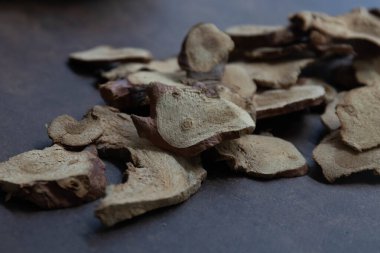 Galangal dilimleriyle dolu siyah bir kase. Ek parçalar koyu dokulu bir yüzeye saçılmış. Topraksı tonlar ve organik şekiller, geleneksel aşçılık ya da yemek pişirme gibi ilaçlarda sıklıkla kullanılan kurutulmuş otların doğal ve kırsal karakterini vurgular.