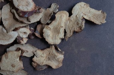 Galangal dilimleriyle dolu siyah bir kase. Ek parçalar koyu dokulu bir yüzeye saçılmış. Topraksı tonlar ve organik şekiller, geleneksel aşçılık ya da yemek pişirme gibi ilaçlarda sıklıkla kullanılan kurutulmuş otların doğal ve kırsal karakterini vurgular.