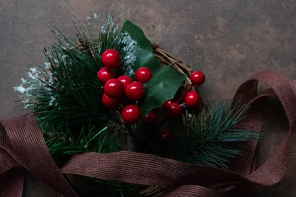 Parlak kırmızı böğürtlenlerle süslenmiş şenlikli çelenk, yapay çam filizleri sahte kar ve yeşil yapraklarla kaplanmış. Kırsal bir kahverengi kurdele düzenlemenin etrafına ustalıkla örtülmüş, karanlık bir metne karşı basit ama büyüleyici bir Noel süsü oluşturuyor.