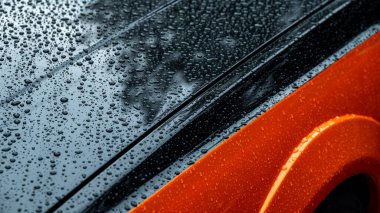 Yüzeyinde yağmur damlacıkları sergilenen turuncu bir arabanın yakından görüntüsü. Yansımalar ve bulanıklıklar sanatsal bir görünüm yaratır, yağmurlu bir günde su ve renklerin etkileşimini yakalar..